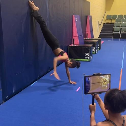 Two young girls in a gym using a tumbling assistant tablet during a Handstand drill.